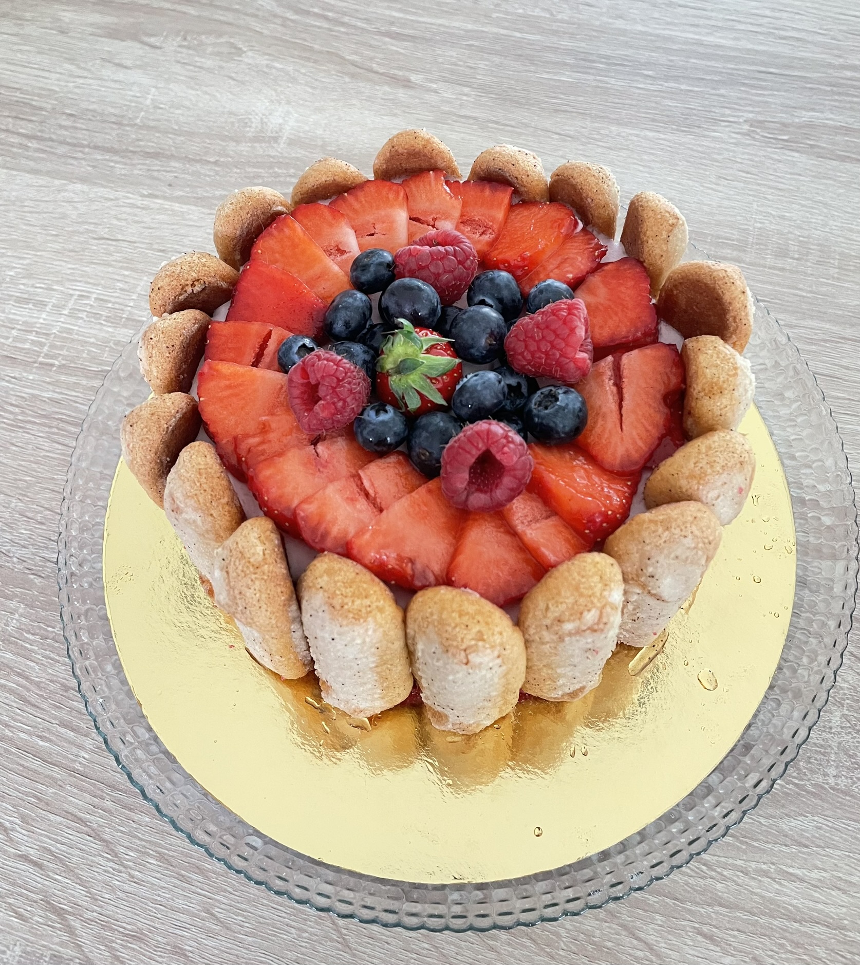 Charlotte aux fraises entourée de biscuits à la cuillère, décorée de tranches de fraises, de framboises et de myrtilles, sur un plat en verre.