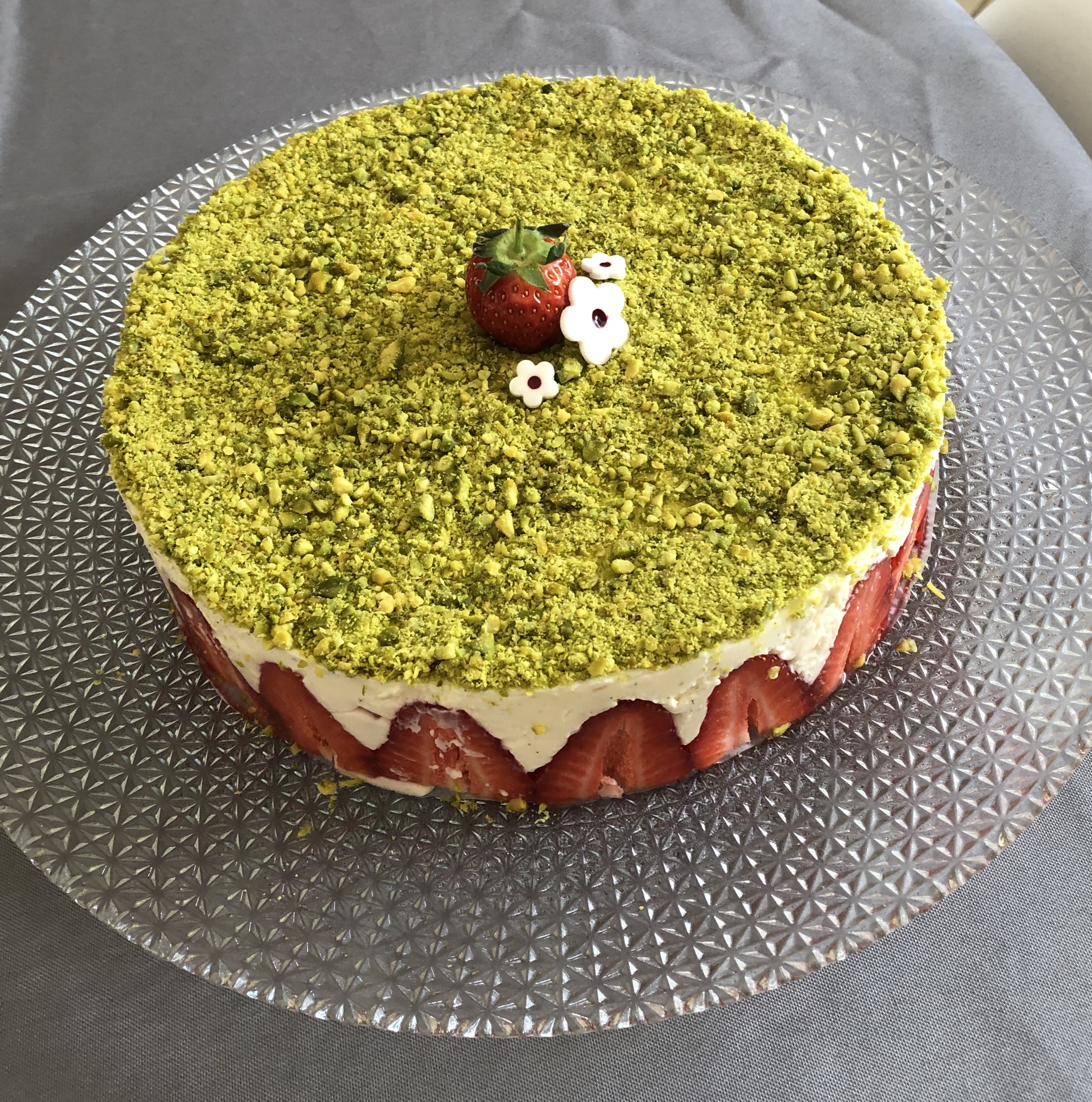 Fraisier à la crème diplomate recouvert de pistaches mixées, décoré d'une fraise et de petites fleurs blanches, sur un plat de service.