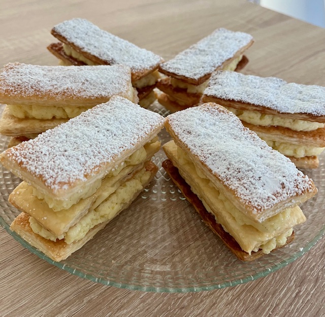 Millefeuilles à la vanille individuels saupoudrés de sucre glace, avec une crème pâtissière entre trois couches de pâte feuilletée caramélisée, sur une assiette en verre