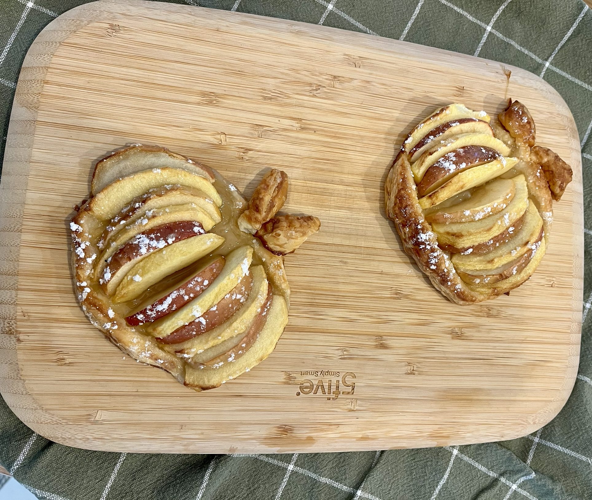 Deux tartelettes fines aux pommes en forme de pomme, avec des lamelles de pommes et une tige en pâte feuilletée, sur une planche en bois.